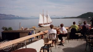 Bilde: Folk på terrassen utenfor Trondenes Historiske Senter. Anleggsbrakker og Anna Rogde i bakgrunnen. Foto: Øystein Normann/Sør-Troms Museum.